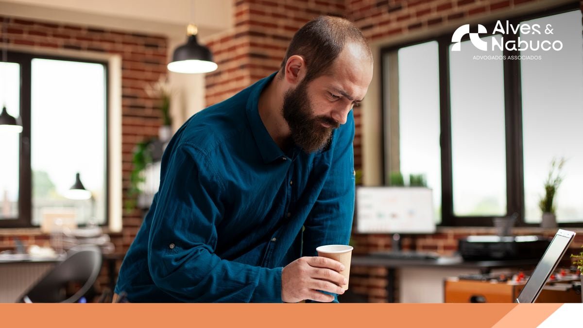 Empresário em escritório, com um copo de café na mão, inclinado sobre a mesa analisando informações no notebook
